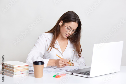 Female blogger wears white shirt, round spectacles, writes notes in notepad, works on laptop computer, drinks coffee, busy with developing webiste, uses wireless interent connection, isolated on white photo