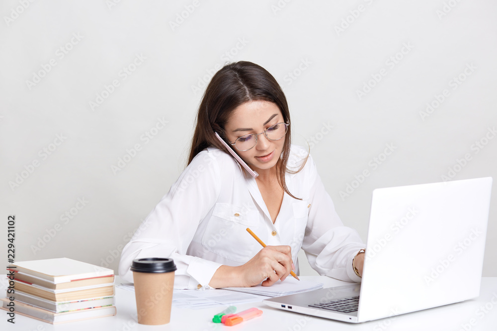 People, technology and business concept. Image of working woman has phone conversation, writes down some notes in notebook, searches information on laptop computer, drinks coffee, isolated on white People, technology and business concept. Image of working woman has phone conversation, writes down some notes in notebook, searches information on laptop computer, drinks coffee, isolated on white