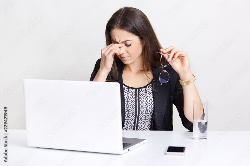 Photo of overworked businesswoman takes off spectacles, dressed in stylish black jacket, works on laptop computer, uses mobile phone, isolated over white studio wall. Tiredness and work concept Photo of overworked businesswoman takes off spectacles, dressed in stylish black jacket, works on laptop computer, uses mobile phone, isolated over white studio wall. Tiredness and work concept