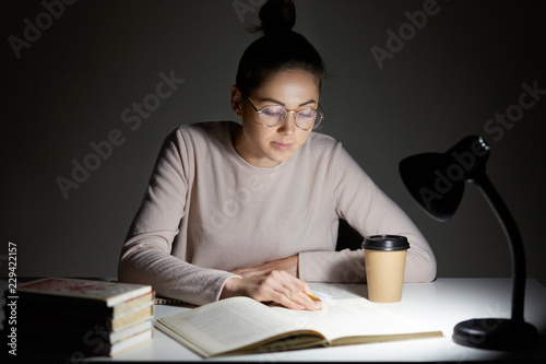 Busy female teenager reads book, uses table lamp, prepares for final examination, sits at desktop, wears optical round glasses, poses against dark background. Reading in evening. Studying concept photo
