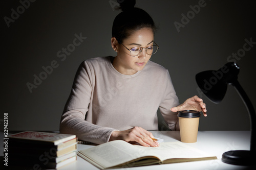 People and exam preparation concept. Attrative young woman dressed in casual clothes, has hair knot, reads scientific literature with cup of coffee, uses reading lamp for good vision in darkness photo