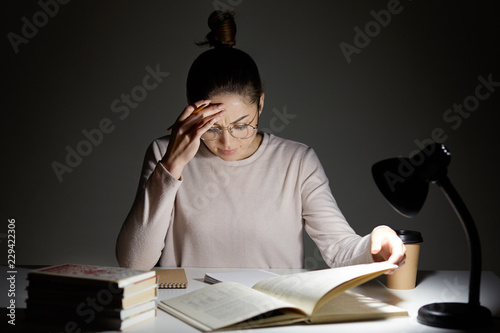 Stressful schoolgirl does home assigment, rewrites information from book in notebook, keeps hand on forehead, feels tired, works late at night, has deadline, dressed in casual outfit round glasses photo