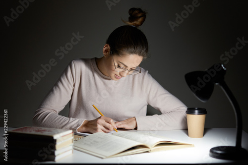 Busy freelancer rewrites infrormation into notepad, prepares article for publication, reads books, writes some notes in organizer, drinks takeaway coffee, sits in darkness, wears round big spectacles photo