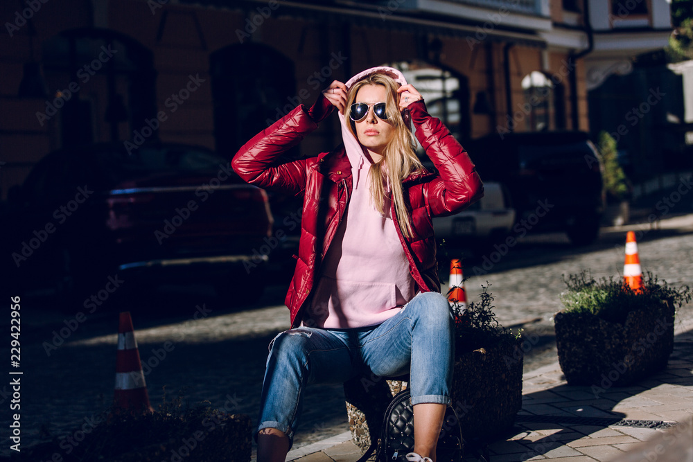 Street fashion concept - portrait of a beautiful girl sitting outside, blue jeans, sunglasses, hard lights, red jacket, autumn weather