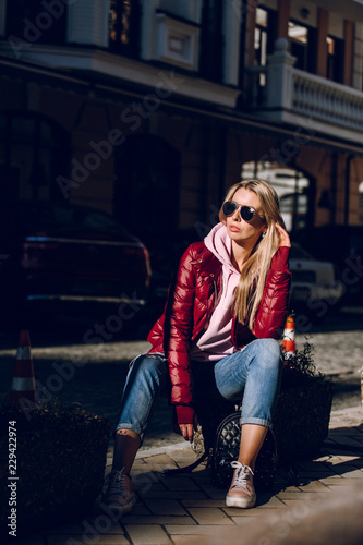 Street fashion concept - portrait of a beautiful girl sitting outside, blue jeans, sunglasses, hard lights, red jacket, autumn weather © boykovi1991