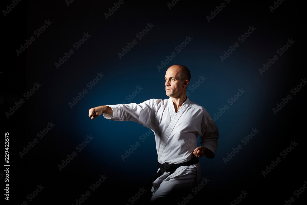 On a blue background with a gradient, an athlete with a black belt does formal karate exercises. On a blue background with a gradient, an athlete with a black belt does formal karate exercises.