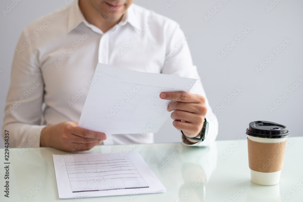 Close-up of smiling young businessman looking at document. Caucasian hr manager sitting at table with coffee cup and reading candidate resume. Employment concept Close-up of smiling young businessman looking at document. Caucasian hr manager sitting at table with coffee cup and reading candidate resume. Employment concept