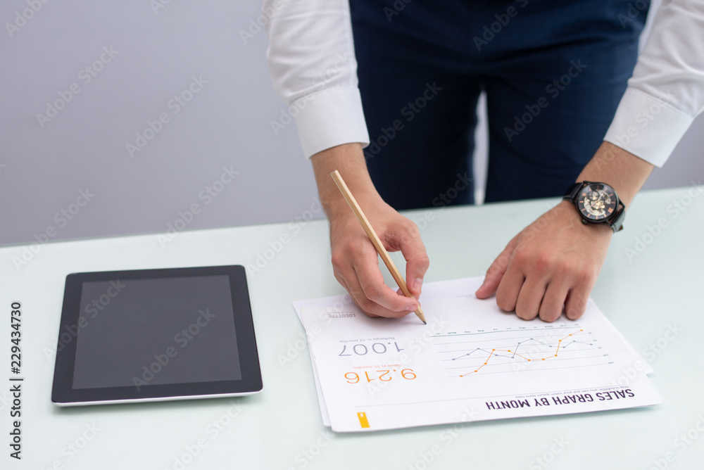 Young marketing manager writing notes on report in office. Caucasian businessman standing at table with digital tablet and working with statistic report. Marketing concept Young marketing manager writing notes on report in office. Caucasian businessman standing at table with digital tablet and working with statistic report. Marketing concept