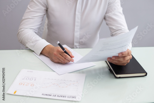 Young businessman sitting at table and working with papers. Caucasian manager sitting at table, looking at document and writing report. Working businessman concept