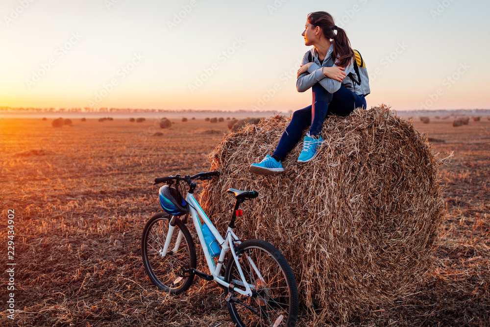 Young bicyclist having rest after a ride in autumn field at sunset. Woman admiring view sitting on haystack - obrazy, fototapety, plakaty 
