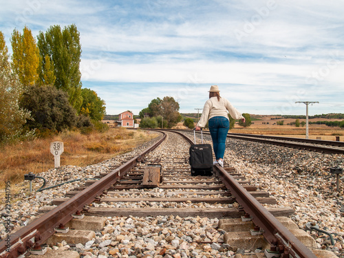 A sad blonde woman with a straw hat walking on the train tracks carrying a suitcase in autumn