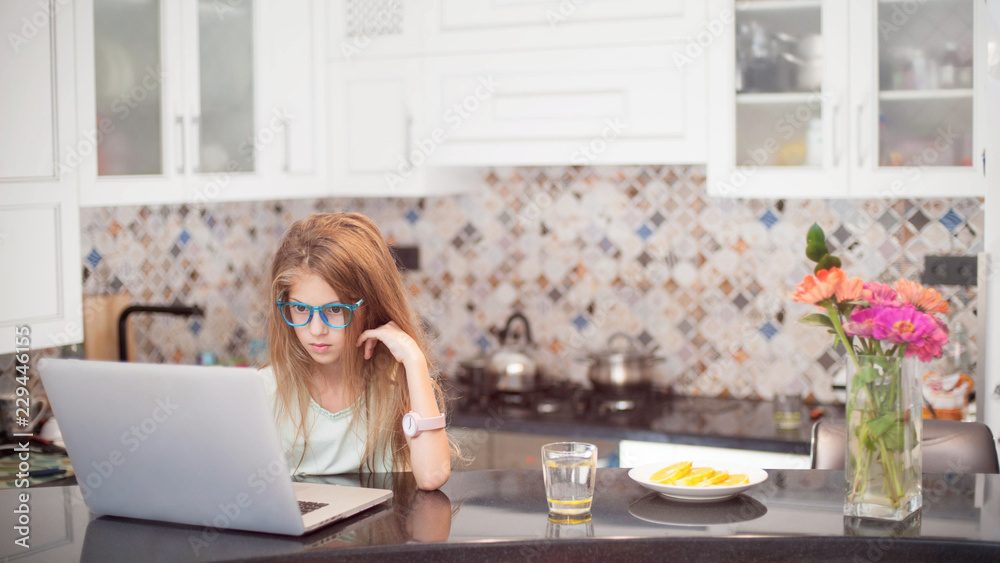 beautiful caucasian little girl in glasses sitting in kitchen using wireless laptop