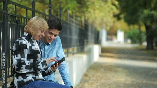  Optimistic view of a cheery blond woman giving high five to her sportive brunet son sitting on a fence parapet and surfing the net in autumn  