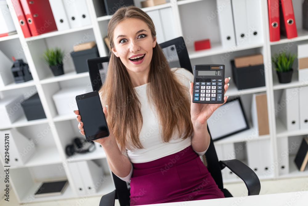 A young girl sitting at a table and holding a phone and a calculator. A young girl sitting at a table and holding a phone and a calculator.