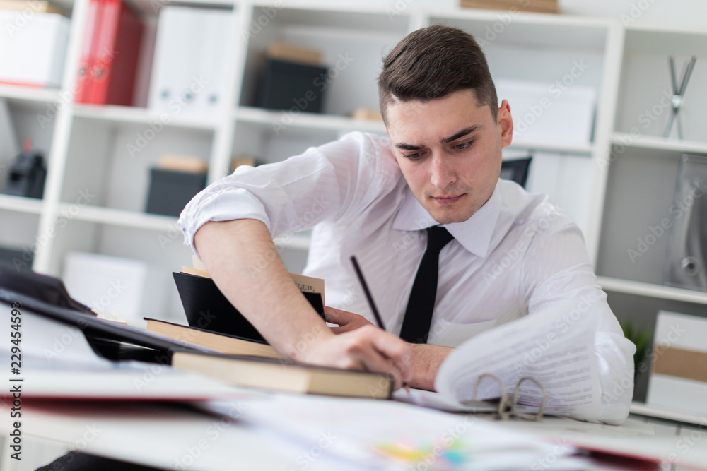 A young man working at a table in the office with a book, documents and a computer. A young man working at a table in the office with a book, documents and a computer.