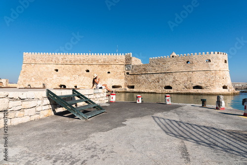 Happy tourist girl on holiday trip to Heraklion, Crete, Greece. Young woman traveller enjoyes sunshine sitting on the dock of Heraklion Venetian port marina. Venetian fort at background. photo