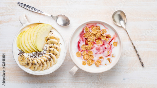 Two greek yogurt with jam, apple, muesli, chia seeds and banana in white bowl on white wooden table, top view, banner