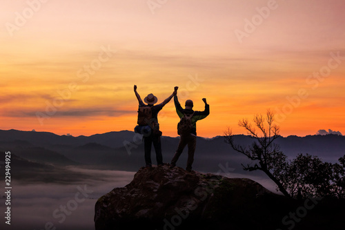 Two tourists with backpacks in the campaign raise their hands up on morning foggy mountain peak. Panoramic view of tourist on mountains hill raising his hands. Successfully achieving your goal concept