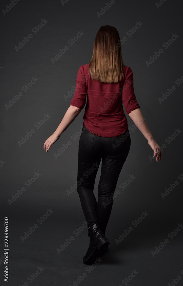 full length portrait of brunette girl wearing red shirt and leather pants. standing pose, facing away from the camera, on grey studio background. full length portrait of brunette girl wearing red shirt and leather pants. standing pose, facing away from the camera, on grey studio background.