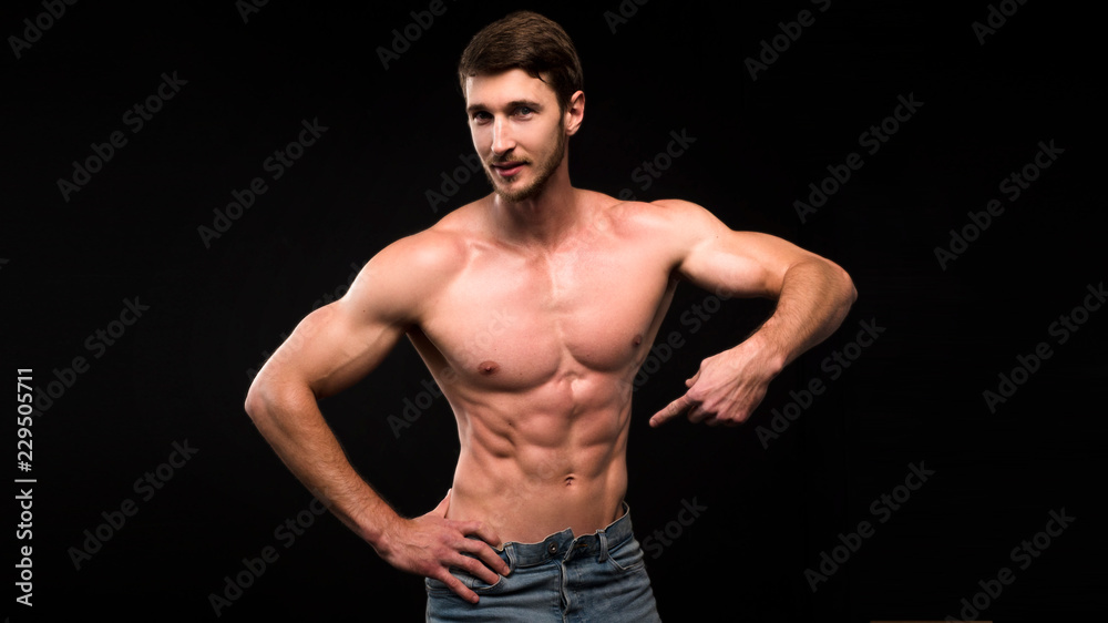 Handsome young muscular man shirtless wearing jeans, on dark background in studio shot. Showing his ABS by finger.