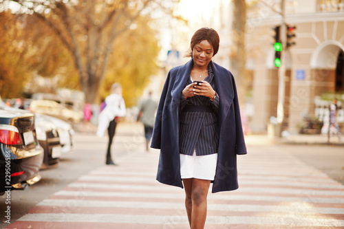 Success stylish african american woman in coat walking at crosswalk and looking on phone. © AS Photo Family