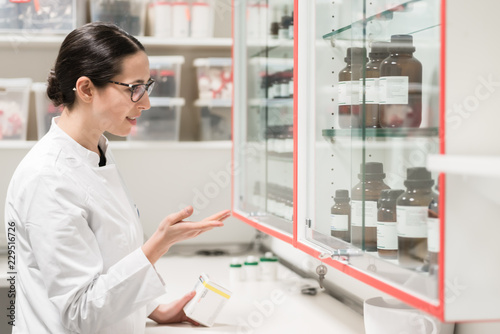 Side view of an experienced female pharmacist checking the container of a chemical pharmaceutical substance during inventory in a modern drugstore