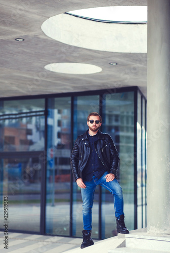 Trendy guy with blue jeans and leather jacket and eyeglasses standing by urban background