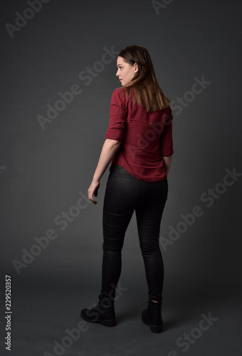 full length portrait of brunette girl wearing  red shirt and leather pants. standing pose, facing away from the camera, on grey studio background.
