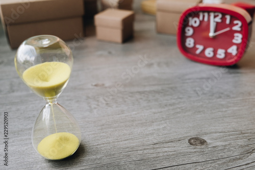 Hourglass as time passing concept with red clock and cardboard box on wooden background.