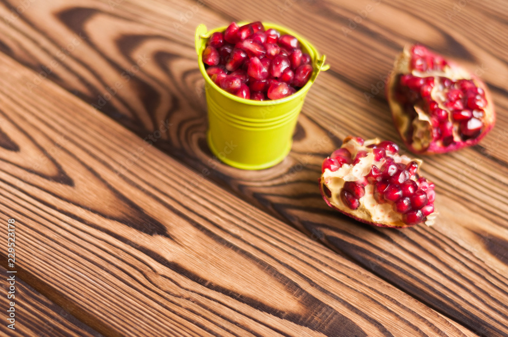 Two halves of fresh ripe red pomegranate with seeds near full green metal bucket of seeds on old brown weathered rustic wooden table Two halves of fresh ripe red pomegranate with seeds near full green metal bucket of seeds on old brown weathered rustic wooden table
