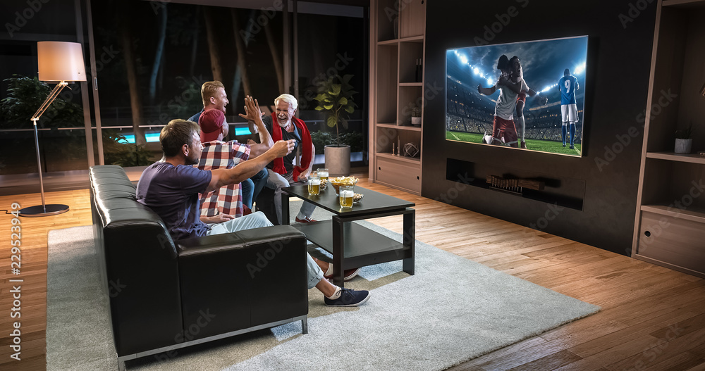 Group of fans are watching a soccer moment on the TV and celebrating a goal, sitting on the couch in the living room.