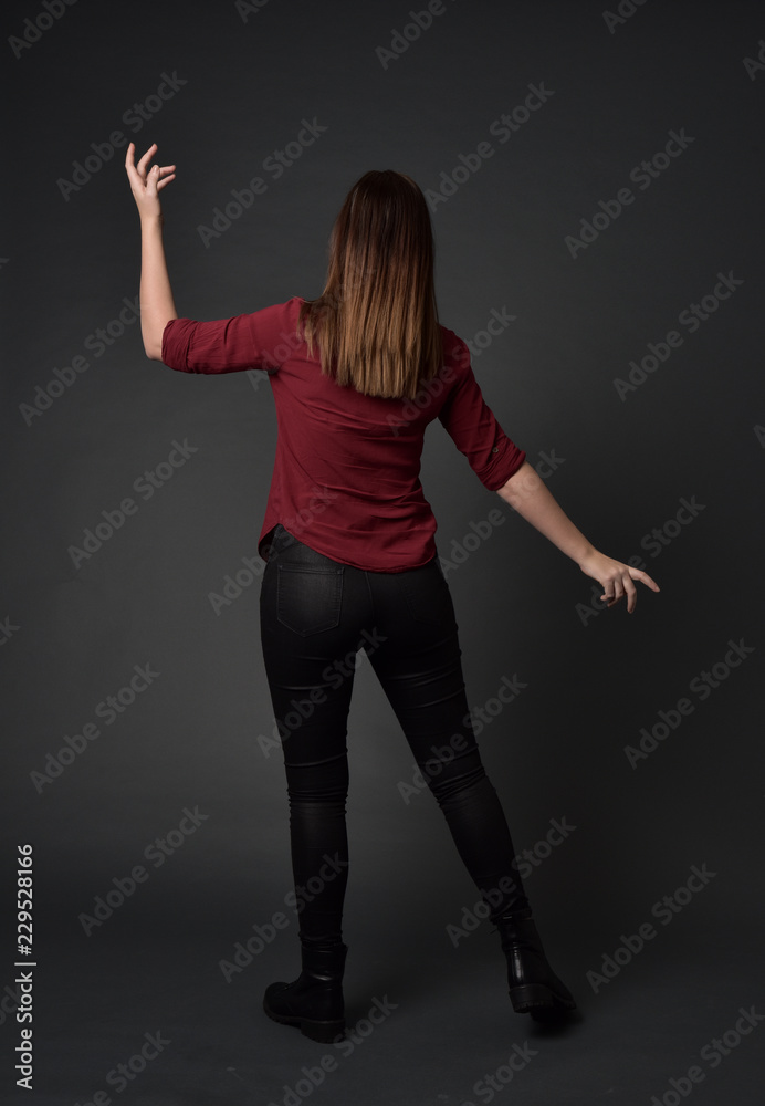 full length portrait of brunette girl wearing  red shirt and leather pants. standing pose, facing away from the camera, on grey studio background.