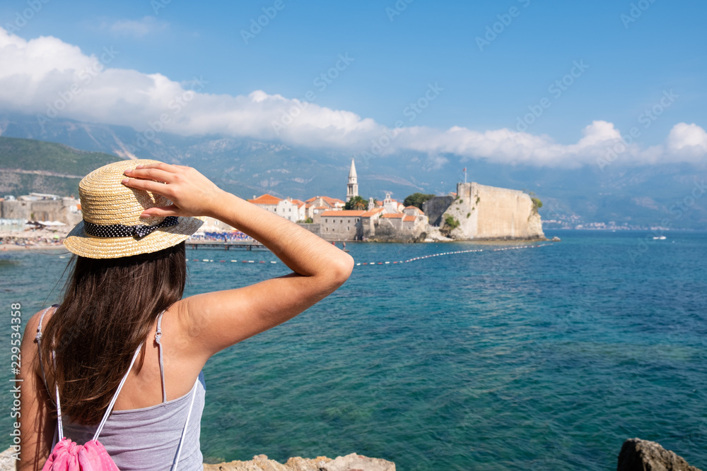 Back view of a young beautiful woman in a hat looking to the old town Budva, Montenegro