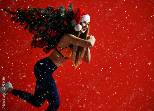 Santa hat Christmas sporting woman holding  xmas tree on her shoulders. Winner energy he red background. it's snowing