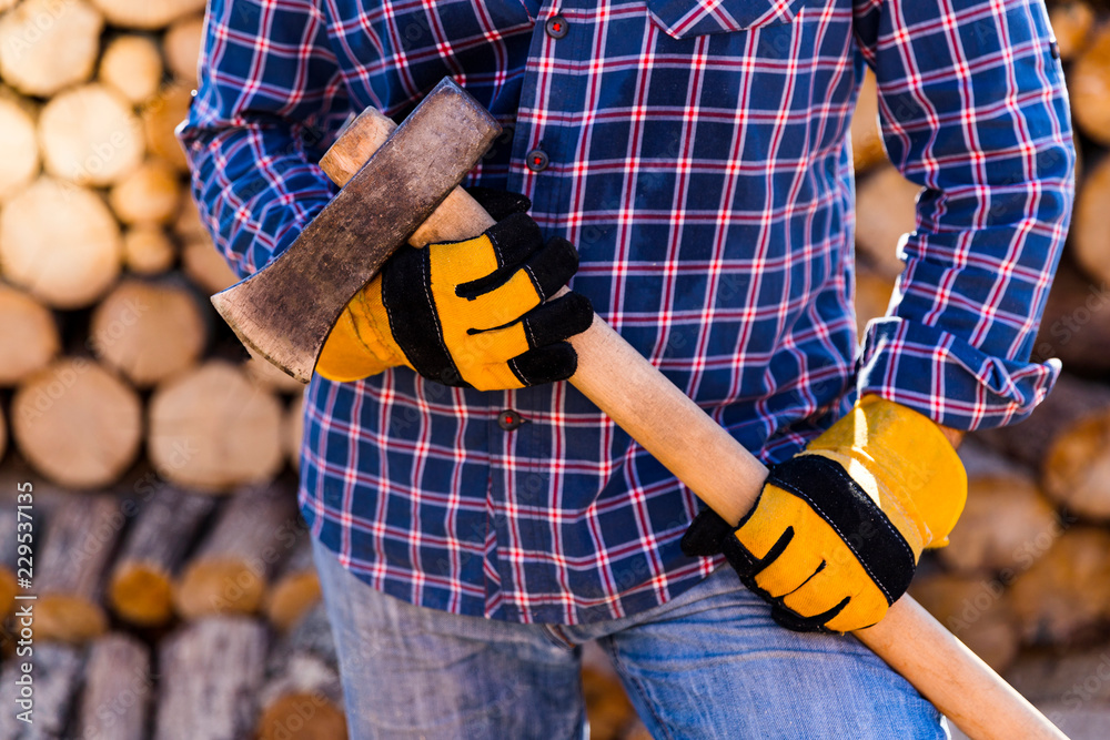 Ax in hand. Man holding an industrial ax. A strong man holds an ax in his hands against the background of firewood. Strong man lumberjack with an ax in his hand.