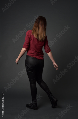 full length portrait of brunette girl wearing red shirt and leather pants. standing pose, facing away from the camera, on grey studio background.