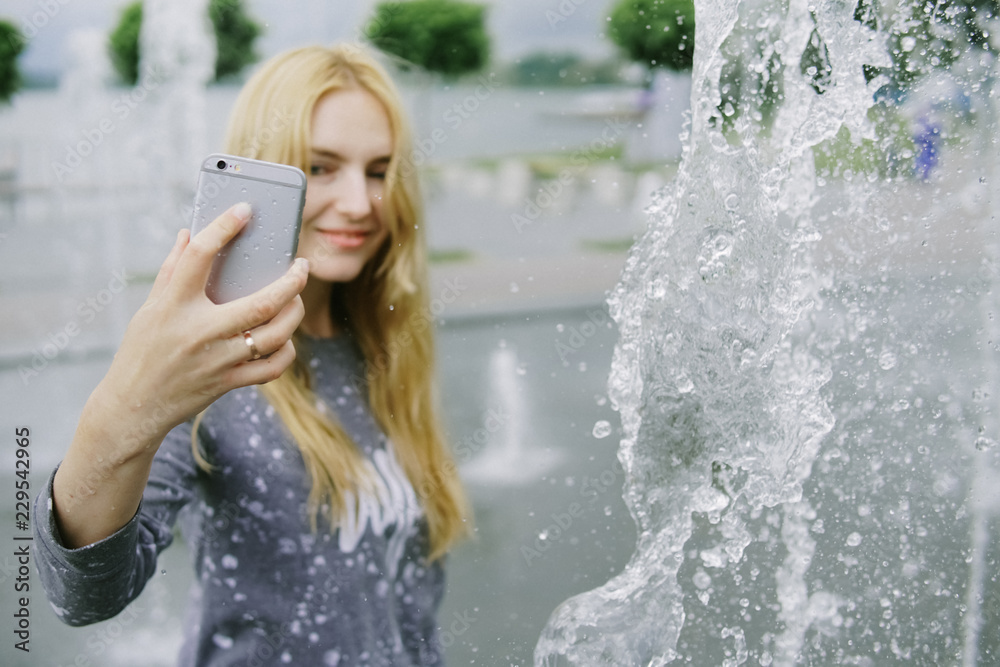 Obraz premium Portrait of beautiful student teenager girl holding up a smartphone taking selfies photos, networking using technology, outdoors park. Adolescent with books near water fountain, lifestyle exterior.