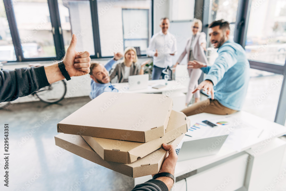 Obraz premium cropped shot of man holding pizza boxes and showing thumb up to colleagues in office