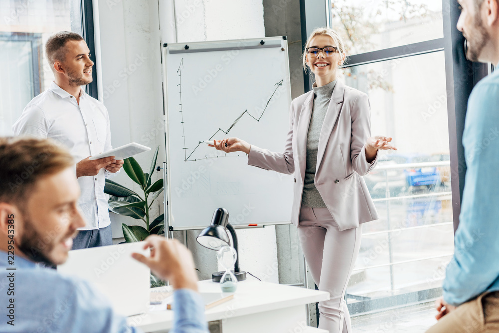 Obraz premium smiling young businesswoman standing near whiteboard and looking at male colleagues in office