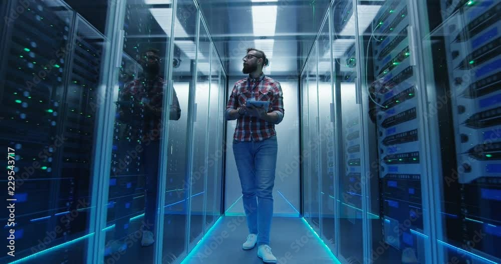 Wide shot of casual man with tablet walking among server racks in data center room looking at hardware