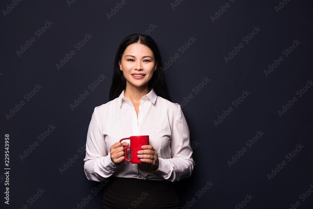 Smiling confident business woman posing on camera and holding red coffee cup. Isolated on dark background. Smiling confident business woman posing on camera and holding red coffee cup. Isolated on dark background.