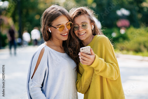 Two funny smiling sisters making selfie on smaptphone and listening music, posing on the street, vacation mood, crazy positive feeling, summer bright clothes sunglasses. photo