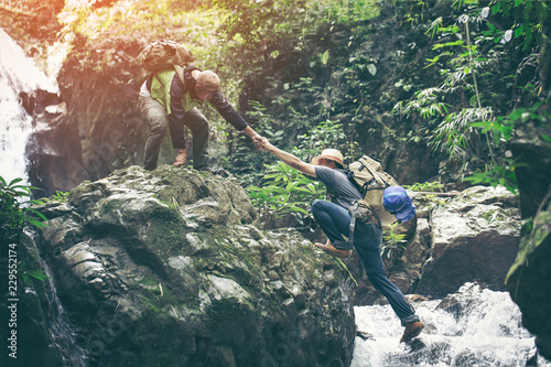 Two Hikers young Man holding hands helping each other walking mountain stream with waterfall
