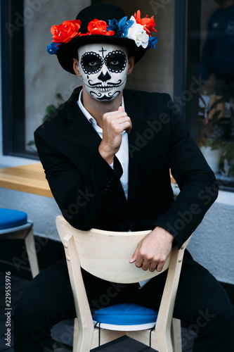 a man with a painted face of a skeleton, a dead zombie, in the city during the day. day of all souls, day of the dead, halloween, ghost walk.