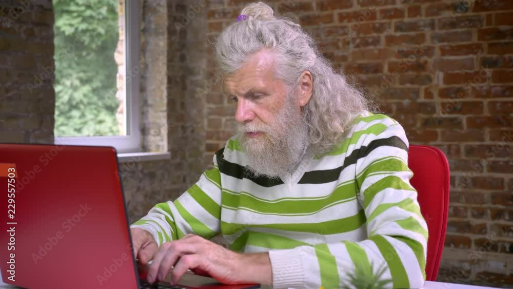 Awesome hairy aged caucasian man with long white beard is typing and demonstrating winning gesture with his fist while sitting at his desktop on brick background Awesome hairy aged caucasian man with long white beard is typing and demonstrating winning gesture with his fist while sitting at his desktop on brick background