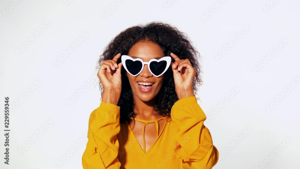 Happy african american woman with afro hair in yellow top wearing heart shaped trendy sunglasses over white wall background. Girl having fun, flirting, smiling to camera.