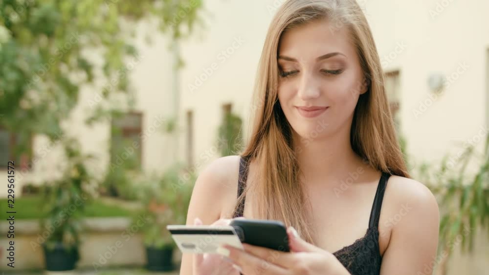 An attractive young lady using a phone and holding a credit card in town. Medium shot. Soft focus An attractive young lady using a phone and holding a credit card in town. Medium shot. Soft focus