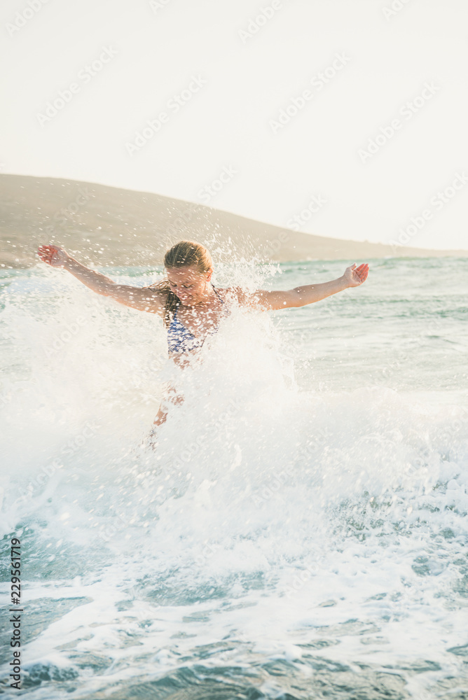 Young beautiful blond woman tourist in swimsuit standing and enjoying splash of wavy waters of Mediterranean sea in Prasonisi cape, Rhodes, Greece on summer day