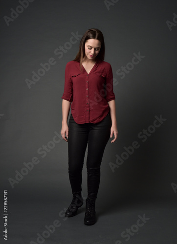 full length portrait of brunette girl wearing red shirt and leather pants. standing pose, on grey studio background.