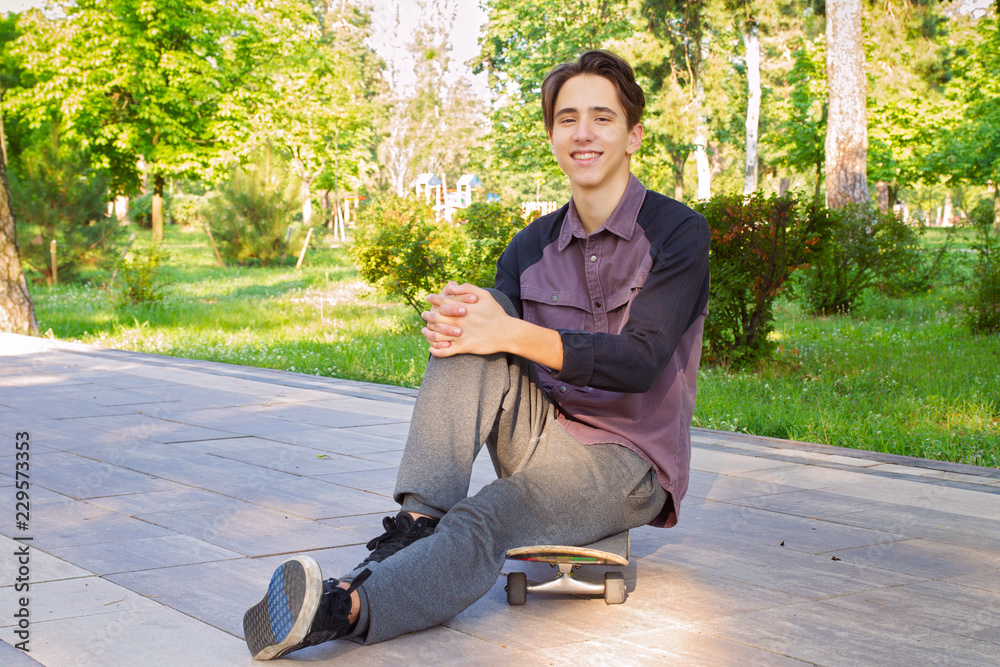 Obraz premium Young man sitting and relaxing on skateboard after skating in city park. Teen boy skater